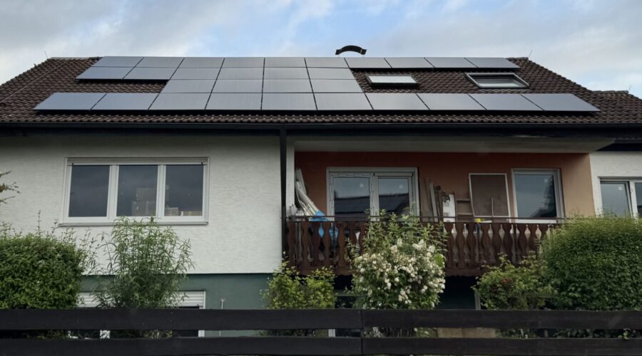 Ein weißes Haus mit einem braunen Dach mit Sonnenkollektoren, umgeben von grünen Büschen und Bäumen, mit einem Holzzaun davor und einem bewölkten Himmel darüber.