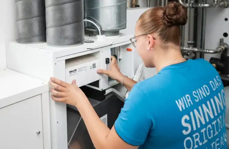 Eine Frau im blauen Hemd bedient oder prüft eine Lüftungs- oder HLK-Anlage, indem sie in einem Technik- oder Maschinenraum die Regler der Anlage einstellt.