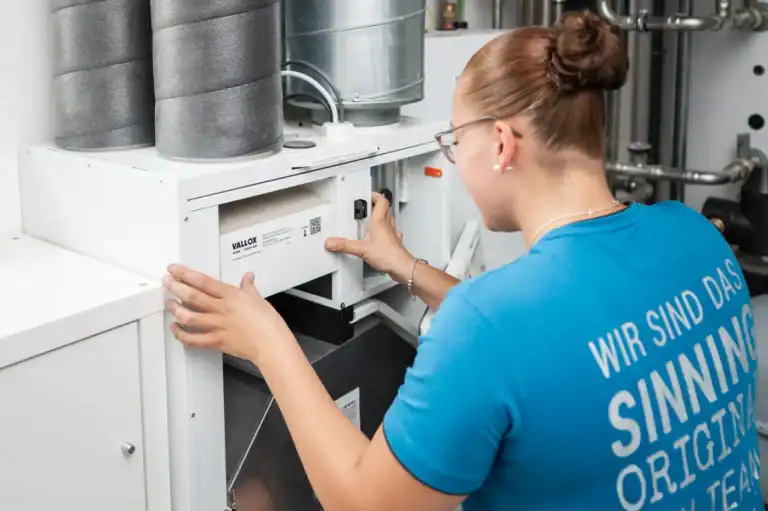 Eine Frau im blauen Hemd bedient oder prüft eine Lüftungs- oder HLK-Anlage, indem sie in einem Technik- oder Maschinenraum die Regler der Anlage einstellt.