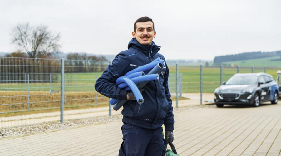 Eine Person in einer marineblauen Jacke steht im Freien auf einer gepflasterten Fläche und lächelt, während sie blaue Rohre über der Schulter trägt und eine grüne Tasche hält. Im Hintergrund sind ein Auto und Felder zu sehen.
