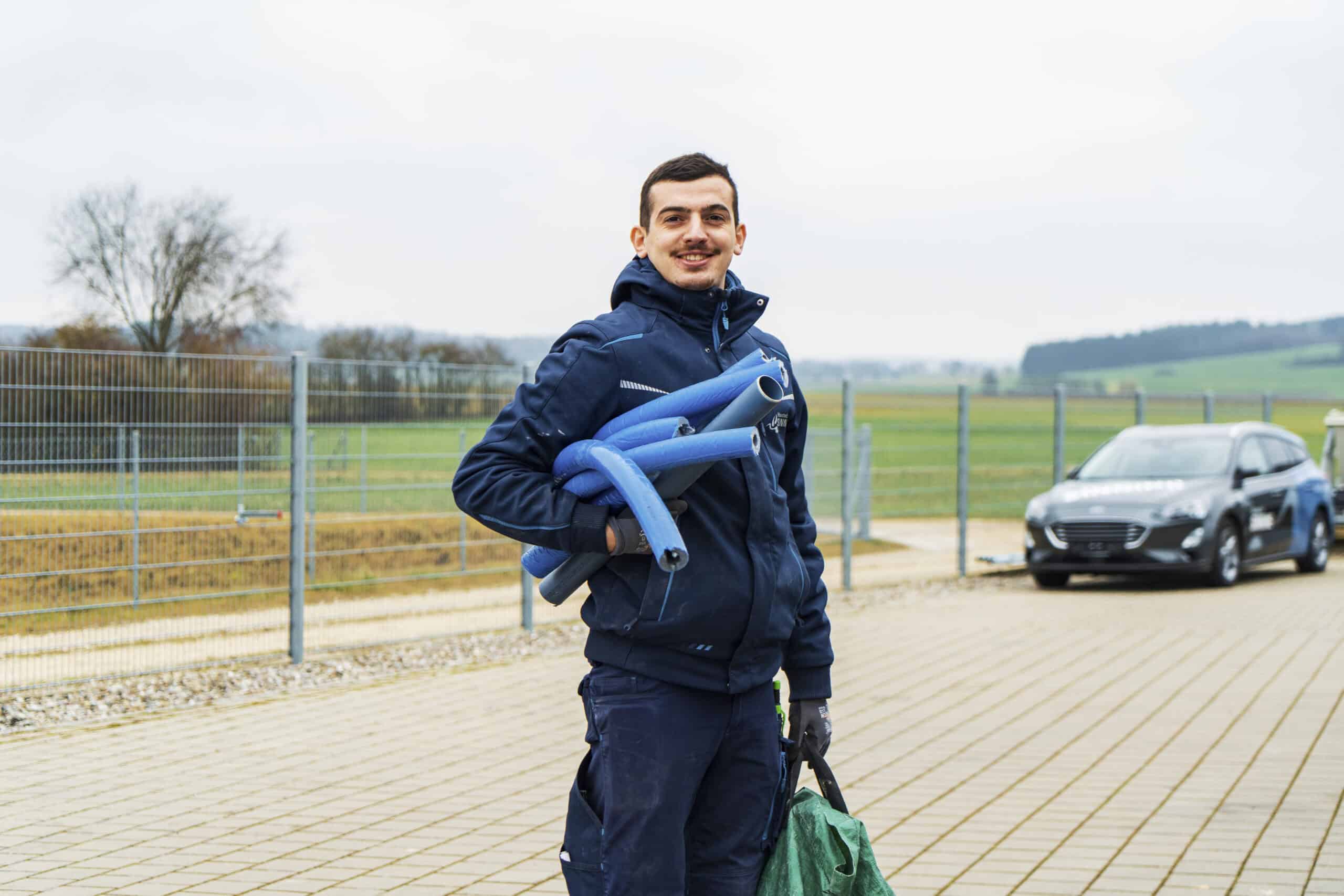 F_SINNING_CP_121225_178 Eine Person in einer marineblauen Jacke steht im Freien auf einer gepflasterten Fläche und lächelt, während sie blaue Rohre über der Schulter trägt und eine grüne Tasche hält. Im Hintergrund sind ein Auto und Felder zu sehen.